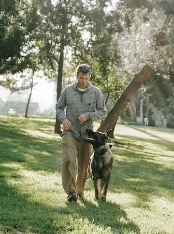 Mike walking with a German Shepherd on a leash through a sunny park with trees and grass for Palos Verdes K9.